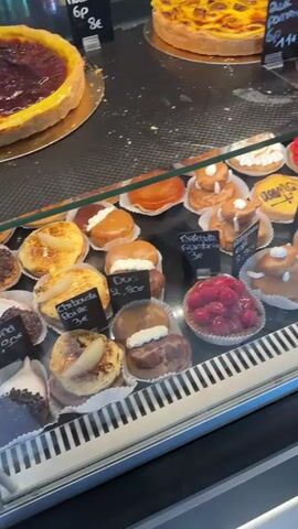 A display case filled with various pastries is shown. There are large tarts on the top shelf, including one with fruit and one with a pear topping. Below are smaller pastries such as donuts, cream puffs, fruit tarts, and chocolate tarts. Several pastries have small price tags attached. A person's hand and arm are visible in the lower right corner of the frame, reaching towards the display. The display case is well-lit, and the pastries appear fresh and colorful. The environment suggests a bakery or pastry shop.