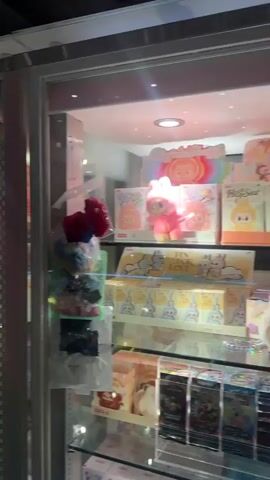 The video shows a display case filled with various plush toys and figurines. The case has glass doors and multiple shelves. Toys visible include plush animals with bunny ears, Stitch figures from "Lilo & Stitch," and various boxed merchandise. The lighting is bright, illuminating the contents of the case. The camera zooms in on a specific white plush toy with bunny ears and a distinctive stitched smile.
