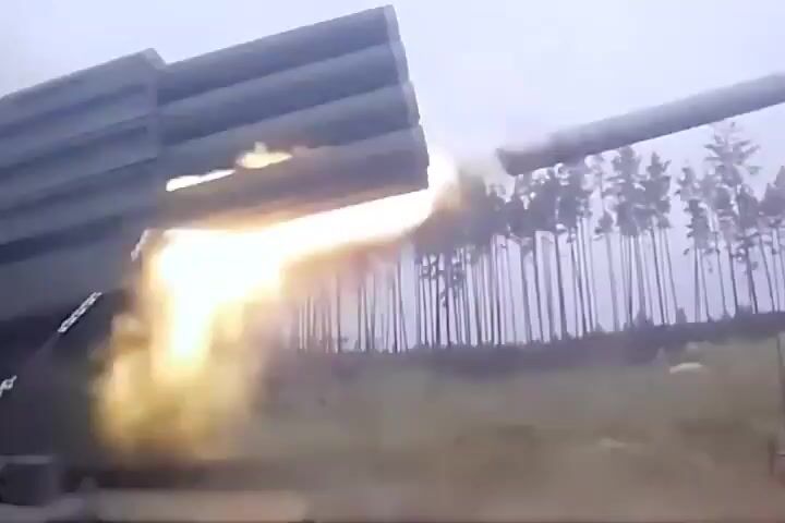 A rocket artillery system mounted on a truck fires several rockets. The rockets are visible as they launch from the tubes and streak through the air. Smoke and dust are kicked up by the firing. In the background, a forest of trees is visible. The video appears to have been filmed from a low angle, looking up at the rocket launcher. The sky is overcast and the lighting is dim, suggesting that it may be dawn or dusk. The overall impression is one of a military operation taking place in a rural setting.