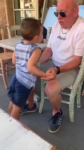 The video shows an elderly man with white hair and sunglasses sitting in a chair, wearing a white t-shirt with an Adidas logo and grey shorts. He is holding hands with a young boy who is standing and facing him. The boy is wearing a blue and white striped tank top and dark blue shorts. They appear to be at an outdoor cafe or restaurant, with tables and chairs visible in the background. The scene is set during the daytime, with sunlight suggesting fair weather. The overall mood seems playful and affectionate, as the man chuckles while interacting with the child. The text visible on the man's shirt is "adidas".