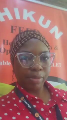 A medium close-up shot of a woman wearing glasses and a red polka-dotted shirt, standing in front of an orange banner. The banner has white lettering that appears to read "HIKUN FEED" and "Healthy Growth & Optimiz..." The woman is also wearing a patterned headscarf and a lanyard with the Olam Agri logo. Her expression is neutral as she looks directly at the camera. The lighting is bright and appears to be coming from above. The background is slightly out of focus, but the banner is clearly visible. The overall impression is that of a promotional event or a trade show.
