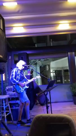 A man with long gray hair is playing a white electric guitar on a stage under blue and yellow lights. He is wearing a blue plaid shirt and grey shorts. To his left is a large speaker and a stool. In front of him are amplifiers, a microphone, and a music stand. In the background, a few people are visible through an open doorway or window. The man is looking down at his guitar as he plays.