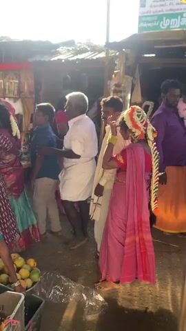 A handheld video shows a lively outdoor market scene. In the foreground, a bright pink sari-clad woman, decorated with flowers in her hair, stands facing left, her head turned slightly as if in conversation. To her left, a man in a white shirt and dhoti looks away from the camera. Behind them, more people are visible, including a woman in a green sari reaching into a crate of fruit, possibly oranges, and other individuals in the bustling marketplace. The background reveals stalls with goods, including framed pictures and garlands, with sunlight streaming in, creating a bright and vibrant atmosphere. The video appears to be shot from behind a metal railing or structure, which partially obscures the view.