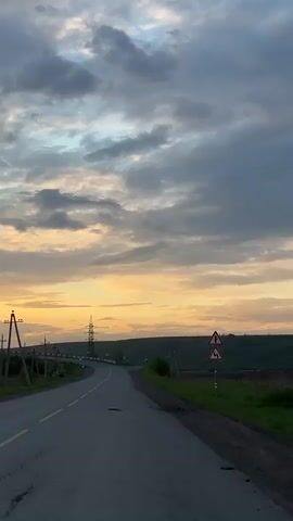 The video shows a view from a moving vehicle on an asphalt road at sunset. The road has a yellow line marking the center and a damaged patch in the lane. On the right side of the road, there's green grass and a road sign indicating a turn, while on the left are fields, trees, and telephone poles with power lines stretching across. In the distance, a bridge and a power transmission tower are visible. The sky is filled with gray and white clouds, with a golden hue from the setting sun illuminating the horizon.