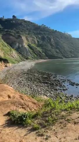 The camera pans from left to right across a coastal scene on a bright, sunny day. The shoreline consists of a rocky beach backed by a steep, verdant hillside covered in trees and vegetation. The ocean is calm, reflecting the sunlight. The sky is blue with wispy clouds, and the sun's glare is visible as the camera pans. Green foliage is visible in the foreground. There are no people visible in the shot.