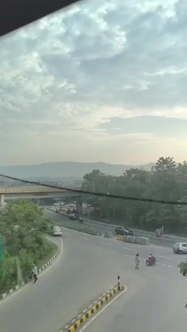 The video shows a view from a moving vehicle looking out at a road and landscape. In the foreground, there is a yellow and black striped barrier. A curved road with multiple lanes is visible, with traffic including cars and a motorcycle. A green signpost with writing in an Indian language is positioned to the left of the road. In the middle ground, a bridge spans across the road, and there are trees and buildings. In the background, hills and mountains can be seen under a cloudy sky. Several people are walking near the road and on a sidewalk. The lighting suggests it is either early morning or late afternoon.