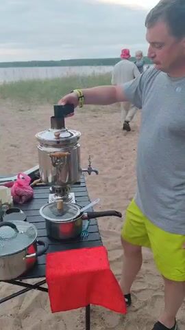 A man with short brown hair and a gray t-shirt is standing on a sandy beach next to a lake. He is holding a black object with a handle and inserting it into the top of a large, silver samovar. The samovar has wooden handles and a spigot on its side. A black table is in front of the samovar with two pots on it, one with a clear lid and another with a black lid and handle. A red cloth is draped over the front of the table. In the background, another person wearing a white top and a pink hat is visible. The sky is overcast.