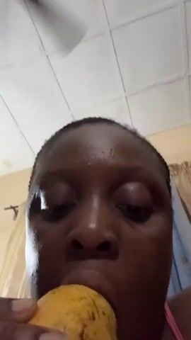 A close-up shot from a low angle shows a dark-skinned woman eating a yellow mango. She is looking down, focusing on the mango in her hand as she takes a bite. Her hair is braided close to her head. The background includes a white, tiled ceiling and a wall with curtains. The lighting is bright, highlighting the sheen on her forehead.