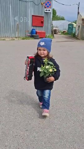 A young girl is standing in a paved area, holding a toy double-decker bus in her left hand and a pot of yellow and white flowers in her right. She is wearing a dark blue beanie with a heart on it, a black hoodie with the word "PUMA" in white letters, blue jeans, and pink sneakers. The area is near a corrugated metal building with a red door and a street sign with a crossing-out symbol and directional arrows. There are also other buildings and some greenery in the background. The sky appears overcast.