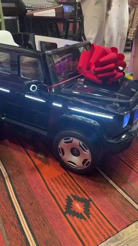 A close-up shot shows a black toy Mercedes-Benz G-Wagon with a large red bow on its hood. The toy car is sitting on a rug with a striped pattern. In the background, several people are partially visible, suggesting a celebratory event. The car's headlights are illuminated with a blue glow.