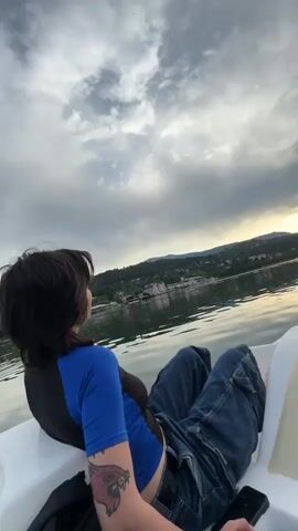 The video begins with a close-up of a person sitting in a pedal boat, looking out at a lake. The person is wearing a blue shirt and jeans, and has a tattoo on their arm. The sky is cloudy, with the sun setting in the distance. The camera then pans to show more of the lake and the surrounding landscape, which includes trees and hills. A sign with text in a foreign language is visible on the boat. The video then cuts to a view from the perspective of the person in the boat, showing their feet and the front of the boat. The sun is visible on the horizon, casting a golden glow on the water. The video ends with a shot of the lake and the sky, with the sun partially obscured by clouds.