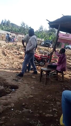 The video shows an outdoor scene, likely in a rural or semi-rural area. The ground is dirt and stone. A red motorcycle is parked under a makeshift shelter or awning. One person is sitting on a small wooden stool next to the motorcycle, appearing to be working on it.  Two men are standing nearby, watching or observing. In the background, there is a building with some signage, several vehicles, and green vegetation. The camera is positioned at an angle and moves slightly.