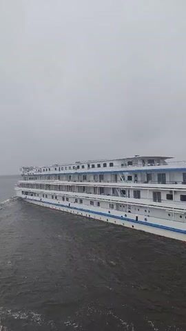 The camera is level and mostly still, pointing towards a large, white, four-story passenger ship navigating a body of water. The ship is moving from left to right in the frame. The sky is overcast and gray. The ship has multiple rows of windows and balconies along its length. Life rafts are visible on the top deck. The water is dark and choppy. The camera appears to be on another boat, as a part of its hull is visible in the lower left corner.