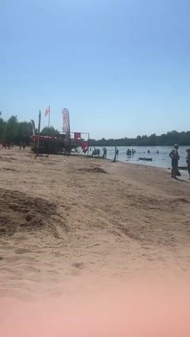 The video shows a sandy beach with the ocean in the background. There are several people on the beach and in the water. On the beach, there are some kayaks and other equipment, along with some flags. The water appears calm, and the sky is clear and blue. Some people are swimming or wading in the water, while others are standing on the shore. The overall scene suggests a recreational day at the beach.