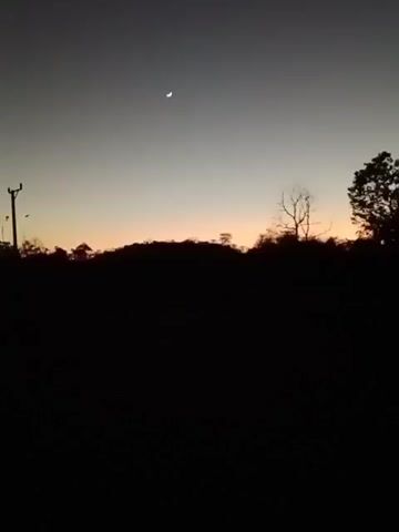The video shows a dark, silhouetted landscape with a crescent moon in the sky at dusk. The foreground is completely black, obscuring any details of the ground. In the middle ground, the silhouettes of trees and hills are visible against a faintly illuminated sky. The sky transitions from a pale orange near the horizon to a dusky gray higher up, with the crescent moon positioned in the upper left quadrant. The overall scene depicts a natural environment at the end of the day.
