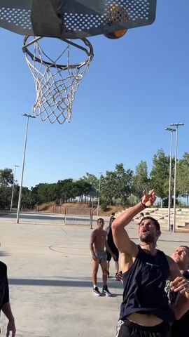 A group of young men are playing basketball on an outdoor court. The camera is looking up at a basketball hoop. In the foreground, a man with a beard wearing a dark tank top is looking up. To his left, a man in a black t-shirt is holding a basketball. Further back, a man with no shirt is looking up, and another man in a black tank top is also looking up. In the background, trees and a clear blue sky are visible. There are streetlights on the court.