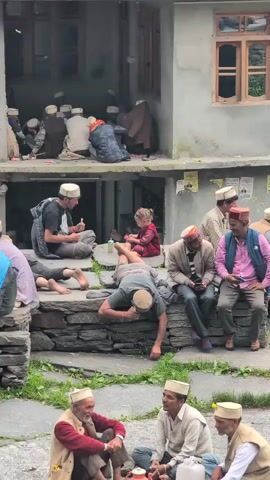 A group of men and one young child are gathered in an outdoor courtyard area. The architecture appears to be of stone and concrete construction, with multiple levels and openings. Several of the men are wearing white caps with red bands. Some men are seated on stone steps and the ground, while others are standing or leaning. One man is playing a musical instrument. The young child, a girl, is seated on the ground and appears to be interacting with something. In the background, on an upper level, more people are visible, some sitting and some standing. The overall scene suggests a social gathering or event.