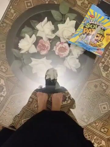 The video shows a person standing with their feet on a patterned rug. The person is wearing a black garment that covers their legs and torso, with the feet visible. The person is reflected in a glass-top coffee table. On the table, there is a bouquet of artificial white and pink roses and a bag of Sour Patch Kids candy. The reflection in the table also shows the person's head and upper body. The video seems to be shot from a low angle, looking up at the person and the coffee table. The surrounding area appears to be a room with patterned flooring.