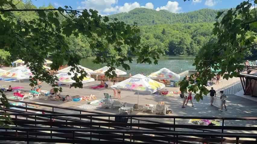 The video captures a sunny day at a lake resort. In the foreground, a black wire fence partially obstructs the view, with green leaves of trees hanging down from above. Beyond the fence, a wooden deck area is set up with numerous beach umbrellas, many of which are white with colorful logos, providing shade for sunbathers. Several lounge chairs and towels are scattered on the deck. People are seen relaxing, sunbathing, and walking around. In the background, the calm lake is visible, reflecting the clear blue sky with fluffy white clouds. Lush green hills covered in dense forest rise behind the lake, creating a picturesque natural setting. A wooden structure, possibly a building or a viewing platform, is situated on the right side of the deck. Further to the left, a path leads away from the deck, with a few people walking on it, and more trees and foliage. The overall atmosphere appears to be one of leisure and recreation on a warm summer day.
