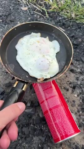 The video captures an outdoor cooking scene set in a natural environment. A person's hand holds a small, black frying pan in which two sunny-side-up fried eggs are cooking. The pan is positioned over a red portable gas canister, partially revealing the word "BUTANE" printed on it, indicating a gas stove setup.

The ground around the cooking area is a mix of dark gravel or charred earth and patches of green grass. As the camera pans upwards, a scenic view of a river or stream comes into focus. The water is a clear, greenish-blue, flowing over a rocky bed. The riverbanks are lined with more grass and gravel, and dense green trees form a lush backdrop across the opposite side, suggesting a wooded or riverside location like Shirokaya Schel, Russia. The overall atmosphere is peaceful, depicting a simple meal being prepared in nature.