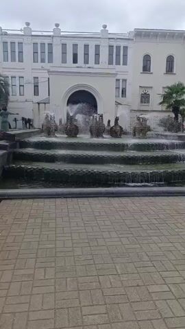A video opens with a fountain in front of a white building. Several tiers of the fountain have water flowing down them, with water spraying up from decorative elements in the center. To the left of the fountain, a person in a green dress stands facing the building with their arms outstretched. Further left, there are palm trees and lush greenery. The ground in front of the fountain is paved with bricks. The camera pans slightly to the left, revealing more of the vegetation and a portion of the white building, including windows and architectural details. The fountain continues to operate throughout the clip.