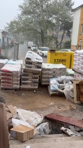 The video shows a construction site with stacks of building materials, including bags of cement and Ytong blocks, under a cloudy sky with heavy rain. Several vehicles are visible in the background, including vans and cars. The ground is wet and muddy, indicating recent or ongoing precipitation. The scene appears to be in an outdoor urban or industrial area. No people are clearly visible in the video.