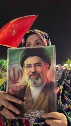 A woman is holding a framed picture of a man in front of her face. The man in the picture has a beard and is wearing glasses, a turban, and traditional Islamic clothing. The picture also has the Iranian flag in the background. The woman is wearing a headscarf and a green robe with a floral pattern. She is standing outdoors at night, with red and green flags visible behind her. The lighting suggests it is an urban or park setting at night.
