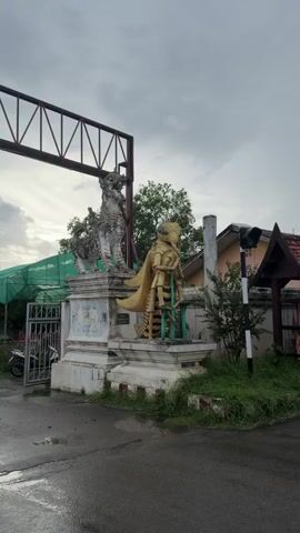 The video shows an outdoor scene with statues and some buildings. In the foreground, there are two prominent statues. The statue on the left is a grey, intricately carved creature resembling a mythical lion or a griffin, standing on a pedestal. To its right is a large, golden statue of a bee-like humanoid figure, also standing on a pedestal. The bee statue has a yellowish body with black stripes, large wings, and appears to be holding a green object. Behind the statues, there is a large, metallic truss structure spanning across the top of the frame. To the left of the statues, a green construction net is visible, with a fence and a gate. Several motorcycles are parked near the gate. In the background, there are trees, a building with a tiered, golden roof that resembles a temple or a traditional Southeast Asian structure, and a traffic light. The ground is wet, indicating recent rain. The sky is overcast with grey clouds. A person wearing a pink jacket is riding a motorcycle on the right side of the frame, moving away from the camera. Another person is visible further down the road. A small food cart with colorful umbrellas is parked near the bee statue.
