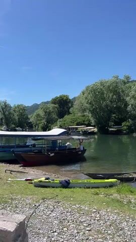 The video shows a wide shot of a lake or river on a clear, sunny day. The water is a deep green, and the shore is lined with gravel and grass. Numerous boats are docked along the shore, including several larger passenger boats and smaller kayaks. The background is filled with lush green trees and rolling hills, under a bright blue sky. There are no people clearly visible or interacting with each other, though some individuals may be on the docked boats.