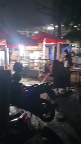 Here is a detailed description of the video:

The video appears to be taken at night in an urban area, likely a street with food vendors or stalls. The initial shots are blurred and shaky, showing the aftermath of what might be a recent event - overturned chairs, scattered debris, and what seems like spilled liquid on the ground. There are figures moving about, some wearing masks, suggesting a possible sense of disorder or cleanup.

As the video progresses, the camera moves, revealing more of the street scene. There are parked motorcycles, and people are seen walking and riding them. The street is lit by streetlights and lights from nearby establishments, and there's moderate traffic with vehicles passing by. In the later shots, some food stalls or mobile vendors are visible, indicating a commercial area. There's a parking sign ("P") visible too. Overall, the scene seems to be a somewhat busy urban street with vendors and people going about their business, despite the initial signs of disarray.