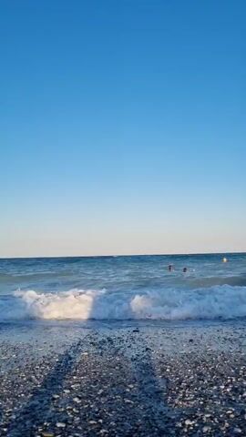 A beach scene under a clear blue sky. Waves crash onto a pebble shore, creating white foam. A few people are visible swimming in the distant water. The sun casts shadows on the beach.