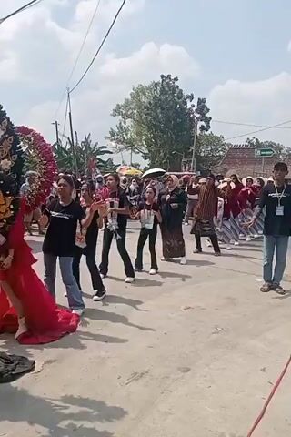 The video shows a procession on a paved road. In the foreground, a person in a red gown with an elaborate headdress stands holding a long golden staff. To their left, another person is visible in dark clothing and a large, blue, feathered headdress. Behind them, a group of individuals dressed in black t-shirts with white sashes are walking and dancing. Further back, a crowd of onlookers watches, some holding umbrellas, and more people are visible in the background, including buildings and trees. The sky is clear and the scene appears to be taking place during the daytime.