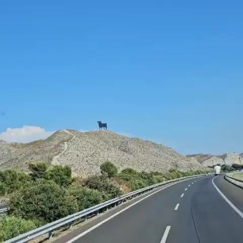 The video shows a landscape view from a moving vehicle on a highway. The sky is clear blue. On the left, a large black bull silhouette stands atop a rocky hill. The highway curves to the right, with white lines marking the lanes. On the right side of the highway, there are various types of vegetation, including trees and bushes. In the distance, a range of hills or mountains is visible. Further along the road, there are several blue road signs. A white truck is seen driving ahead on the highway. Some buildings, including what appears to be a church tower, are visible among the trees in the distance.