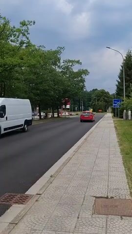 A red car drives down a road past a sidewalk lined with trees and parked cars on the other side. A blue car also drives down the road. The sky is cloudy.