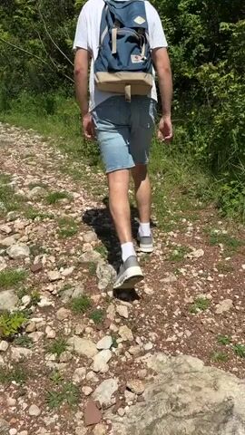 A person walks away from the camera on a rocky path through a wooded area. The person is wearing a white t-shirt, denim shorts, grey shoes with white socks, and a blue backpack with a beige bottom. The path is covered in a mix of dirt, small rocks, and patches of grass. Green foliage and trees line both sides of the path. The lighting suggests it is a bright, sunny day.