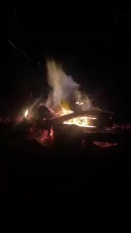 The video shows a campfire burning in the dark. There are logs and embers glowing red and orange. The flames are yellow and white, and there is smoke rising into the air. Sparks fly up from the fire. There are voices heard in the background speaking in French.