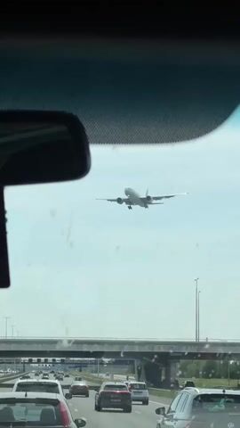 The video shows a car driving on a highway with other vehicles in both directions. Above the highway, an airplane is descending for landing. The video is taken from inside the car, looking through the windshield. The rear-view mirror and a bridge are visible in the foreground.