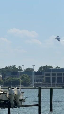 The video shows four Blue Angels fighter jets flying in formation against a slightly cloudy, blue sky. They are leaving white contrails. In the foreground is a dock with a white boat that has two Suzuki outboard motors. A few people are standing on the dock, watching the jets. Behind the dock is a body of water, and across the water is a large building with trees in front of it. There are light poles and other buildings to the sides. The video is shot from a low angle, looking up at the jets.
