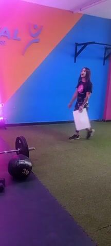 The video, seemingly a short promotional clip, is shot in a brightly lit gym with a colorful wall featuring geometric patterns and the words "INTERVAL Escuela de Fitness" on it. There's a woman with long dark hair wearing a black t-shirt and patterned leggings, holding a white paper, and walking across the room. In the foreground, there is a barbell with weights, a medicine ball labeled "3Kg", and a pair of small dumbbells on a black mat. The floor transitions from black mat to green artificial turf. Spotlights cast pink, purple, and blue hues across the scene. There is also some clapping, possibly in response to the woman being presented.
