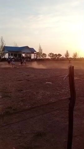 The video shows an outdoor sports field at sunset. Several people are playing soccer. A building with a blue roof is visible in the background, with spectators gathered in front of it. The field is dirt, and dust is kicked up by the players. The sun is low in the sky, casting a warm glow. There is a fence in the foreground.