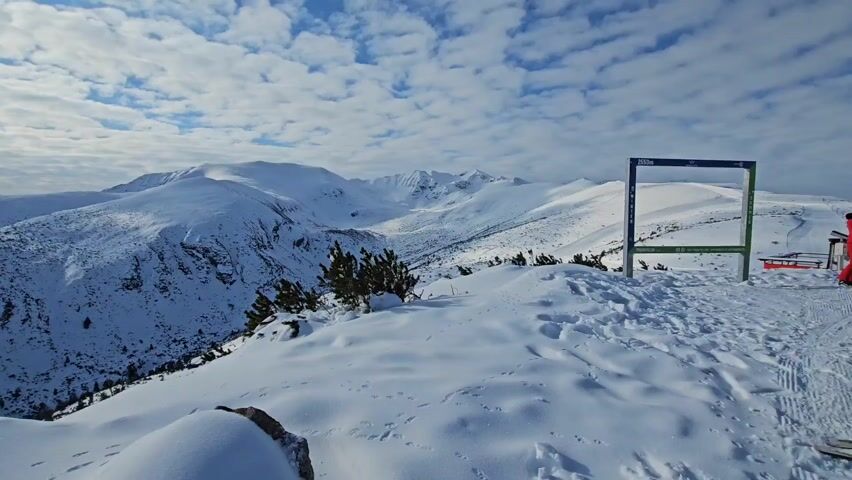 The video showcases a winter scene in the mountains.  The landscape is covered in snow, with rolling hills and mountain peaks visible in the distance.  The sky is a vibrant blue with scattered white clouds, and the sun shines through, creating a bright and airy atmosphere. In the initial frame, there is a green and blue frame, possibly a photo opportunity marker, indicating the elevation.  As the camera pans, the scene expands to reveal a vast expanse of snow-covered terrain.  A sea of clouds is visible in the distance.  Finally, the camera shows a woman in red and white on the left side of the shot, standing by a wooden fence or railing. The overall impression is one of a serene and majestic winter landscape.