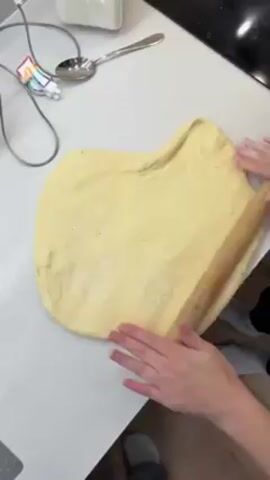 A person is rolling out a piece of dough on a white countertop. They are using a rolling pin and their hands to flatten and shape the dough. A spoon and other kitchen utensils are on the counter nearby. The person is wearing a light-colored sweater.