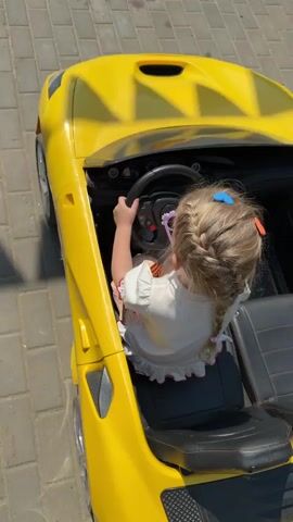 A young girl with braided blonde hair, adorned with blue and orange barrettes, sits in the driver's seat of a yellow, open-top toy car on a paved surface. She is dressed in a white shirt and is holding onto the steering wheel, as if driving. The car has black seats and silver rims on the tires. The pavement beneath is a light grey and consists of rectangular brick-like stones. The shot is taken from above, focusing on the girl and the car.