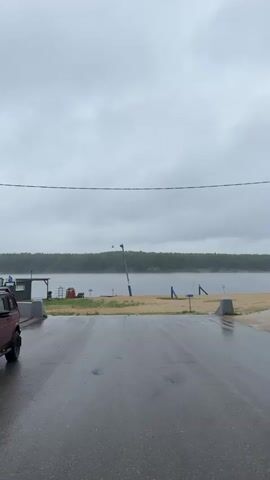 A red SUV is parked on the left side of a wet asphalt road. The road leads to a sandy beach and a body of water. The sky is overcast and appears to be cloudy. There is a line of trees in the background.