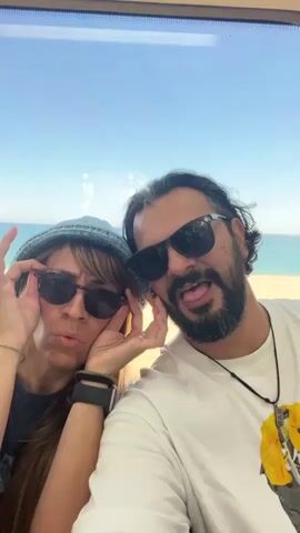 A selfie shows two people wearing sunglasses, with a beach in the background. On the left is a woman with long brown hair and bangs, wearing a blue bucket hat and a black smartwatch. She is holding her sunglasses up, as if adjusting them. On the right is a man with a beard and long dark hair, also wearing sunglasses. He is smiling slightly. Both are wearing casual t-shirts. The background shows a beach and ocean under a blue sky, visible through a window. The location appears to be indoors, possibly a train or building with a scenic view. The selfie is taken at a close range, focusing on the faces of the two individuals.