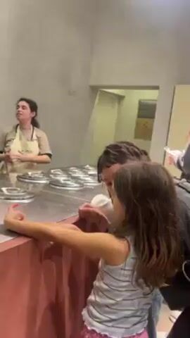A medium shot shows a young girl in a gray and white striped tank top standing at a counter with a person who has dark dreadlocks. They are looking towards a woman standing behind the counter. The woman wears a beige apron and appears to be speaking to the pair at the counter. The countertop has numerous metal lids on it, presumably covering containers below. A sign for "WC" is on the wall in the background, with a partially open doorway visible to the right. Someone holds up a phone or camera in the background.