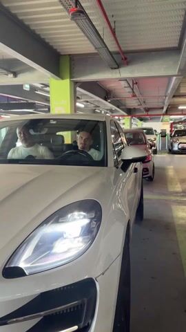 A white Porsche Macan is parked in an indoor parking structure. Inside the car, a young man is seated in the driver's seat, and an older man with white hair is in the passenger seat. The car's headlights are on. The car starts, the sound of the engine briefly audible. The parking structure has green-painted support columns and hanging metal cable trays. Several other cars, including a red one and a white Mini Cooper, are parked further down the row. A green sign reading "ALLEE" is visible on one of the support columns.