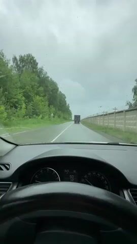 The video shows a first-person view from inside a car driving on a paved road. The dashboard of the car is visible, including the steering wheel, speedometer, and tachometer. The speedometer indicates a speed of 90. The road ahead is empty except for a dark-colored truck in the distance. On the left side of the road, there is a dense line of green trees. On the right side of the road, there is a concrete wall with barbed wire on top. The sky is overcast and appears to be raining lightly, as evidenced by raindrops on the windshield. The overall environment suggests a rural or semi-rural setting.