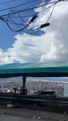 The video shows a cityscape from a high vantage point, possibly a roadside overlook.  The sky is bright blue with puffy white clouds. In the foreground, there's a green canopy tent and a metal guardrail. People are visible under the canopy, and a police presence, including two officers and two green motorcycles, is also noted near the tent. Wires are strung across the top of the frame. The camera appears to be in motion, possibly from a passing vehicle.