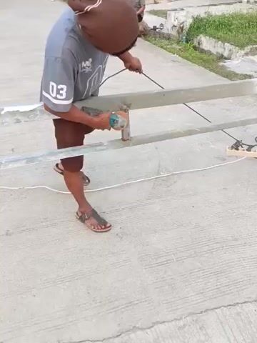 A person wearing a brown beanie, a gray t-shirt with the number 03 on it, and brown shorts is using an angle grinder on a metal pole. The pole is part of a larger metal structure, possibly a fence or railing. The person is standing on a concrete surface, and there is a power cord connected to the angle grinder. Another person is visible in the background, sitting down. The environment appears to be outdoors, with some vegetation in the background.